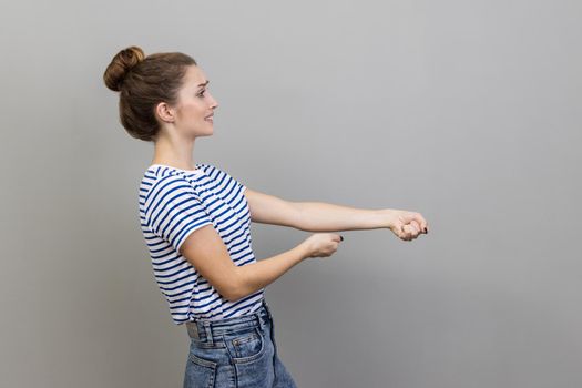 Portrait of strong ambitious woman wearing T-shirt pretending to pull invisible rope, concept of hard working, striving and efforts to achievements. Indoor studio shot isolated on gray background.
