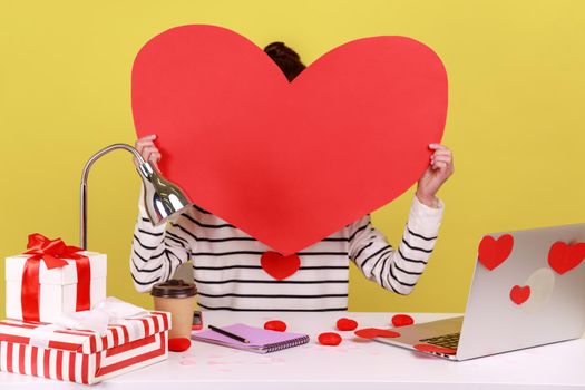 Woman office worker hiding, holding in hands big red paper heart, preparing decoration and gift for saint valentine day celebration. Indoor studio studio shot isolated on yellow background.