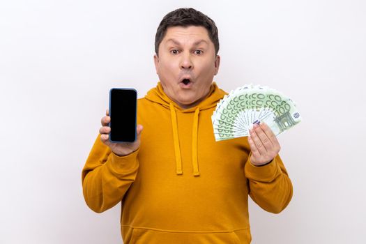 Astonished surprised man holding empty screen smartphone ad fan of hundred euro, banking and earning online, wearing urban style hoodie. Indoor studio shot isolated on white background.