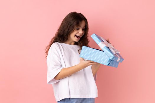 Portrait of little girl wearing white T-shirt opening gift box and peeping inside with excited facial expression, satisfied with present. Indoor studio shot isolated on pink background.