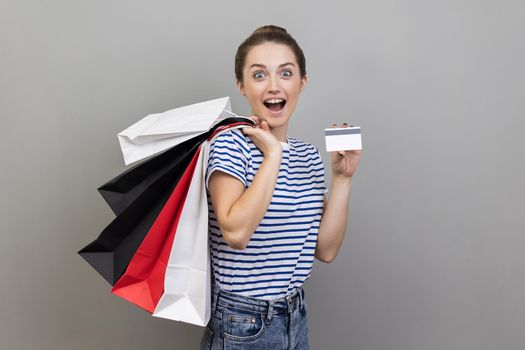 Wow, shopping loan and cashback. Portrait of astonished woman wearing striped T-shirt holding credit card and packages, keeps mouth open. Indoor studio shot isolated on gray background.