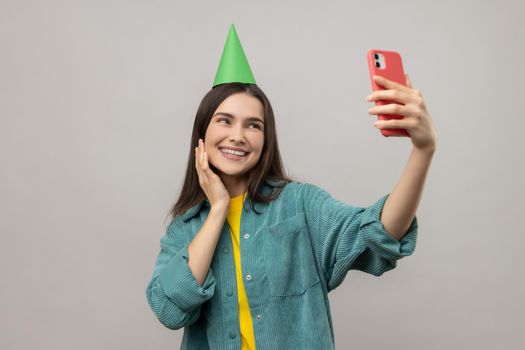 Portrait of cute charming in party cone making selfie or broadcasting livestream, smiling to device camera, wearing casual style jacket. Indoor studio shot isolated on gray background.
