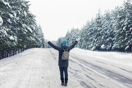 a girl in a gray coat , with a gray backpack on her back, walks along the side of a winter road, raised her hands up. Nearby there is a beautiful winter pines forest. Back view