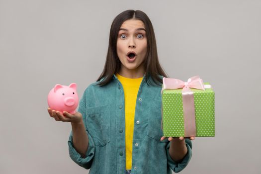 Portrait of amazed surprised woman holding wrapped gift box and piggy bank, save money, looking at camera with open mouth, wearing casual style jacket. Indoor studio shot isolated on gray background.