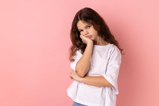 Portrait of lazy bored little girl wearing white T-shirt leaning on hand and looking depressed, child with indifferent gloomy expression has no energy. Indoor studio shot isolated on pink background.