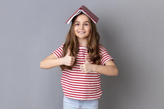 Portrait of delighted little girl in striped T-shirt covering head with book and smiling to camera, child having fun, showing thumbs up. Indoor studio shot isolated on gray background.