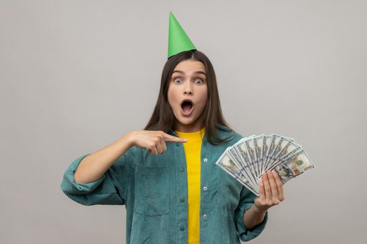 Amazed excited rich woman in party cone on head pointing at money, looking surprised by lottery win, celebrating big profit, wearing casual style jacket. Indoor studio shot isolated on gray background