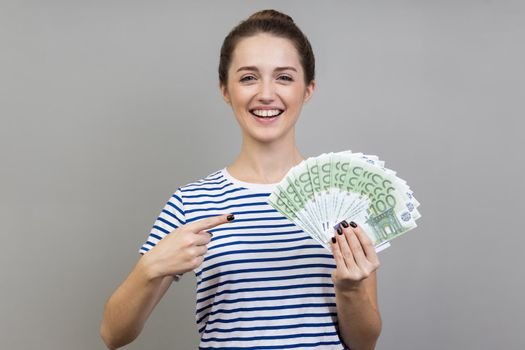 Portrait of woman wearing striped T-shirt pointing finger at fan of euros in her hand looking at camera with toothy smile, cash, money exchange service. Indoor studio shot isolated on gray background.