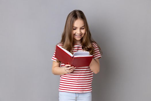 Portrait of smiling satisfied little girl wearing striped T-shirt holds book, enjoys reading, standing absorbed with interesting plot. Indoor studio shot isolated on gray background.