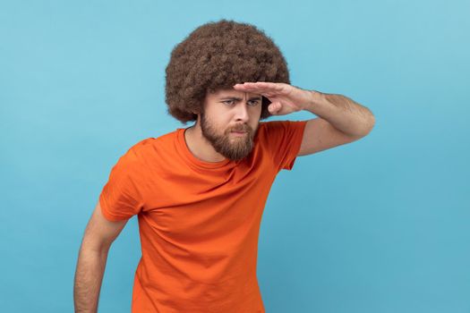 Portrait of man with Afro hairstyle wearing orange T-shirt looking far away at distance with hand over head, attentively searching for bright future. Indoor studio shot isolated on blue background.