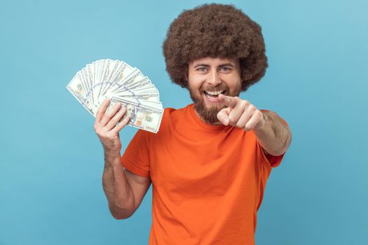 Portrait of smiling happy man with Afro hairstyle wearing orange T-shirt holding dollar banknotes, pointing to camera, you win lottery. Indoor studio shot isolated on blue background.