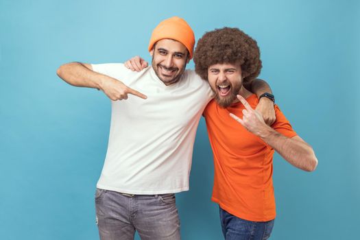 Portrait of two men hugging each other, guy with Afro hairstyle showing rock and roll sign hand gesture, male in beanie hat pointing at his friend. Indoor studio shot isolated on blue background.