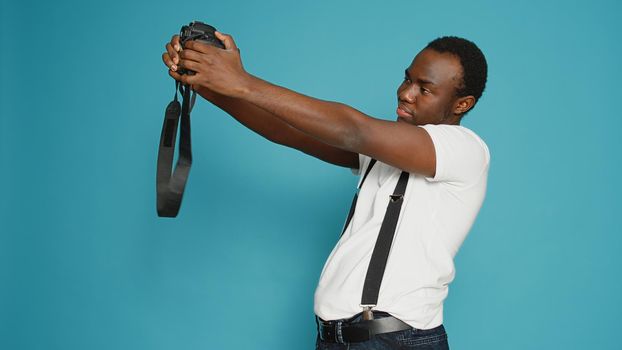 African american person holding dslr camera to take pictures, standing in studio over blue background. Photographer using professional equipment to capture photos, zoom focus snap.