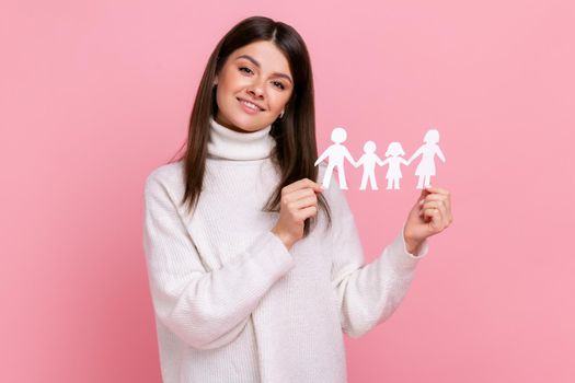 Smiling positive girl holds white paper family chain, looking at camera with happy facial expression, wearing white casual style sweater. Indoor studio shot isolated on pink background.