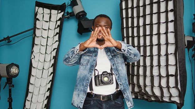 African american photographer doing heart shape symbol with hands, psoing with romantic love gesture over background with photography lighting equipment. Valentines day feelings sign.