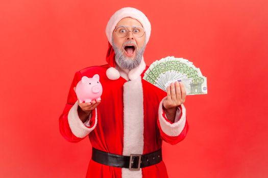 Excited shocked elderly man with gray beard wearing santa claus costume standing with euro banknotes and piggy bank, big amount of money. Indoor studio shot isolated on red background.