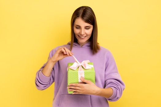 Young beautiful smiling woman feeling happy, opens present box with happy satisfied expression, being glad to get gift, wearing purple hoodie. Indoor studio shot isolated on yellow background.