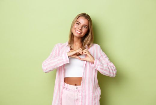 Portrait of beautiful lovely woman with blond short hairstyle, wearing pink outfit, showing heart sign and smiling, confess in love or sympathy, standing over green background.