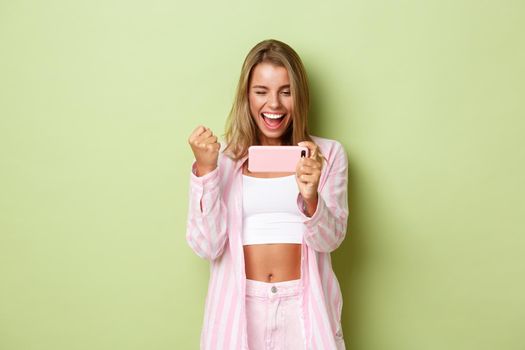 Portrait of happy winning girl with blond hair, looking at smartphone screen and triumphing, standing over green background.