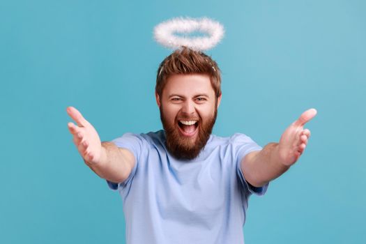 Portrait of excited angelic man with holy nimbus on head standing opening his hands wide to hug, welcoming with friendly smile, sharing love. Indoor studio shot isolated on blue background.