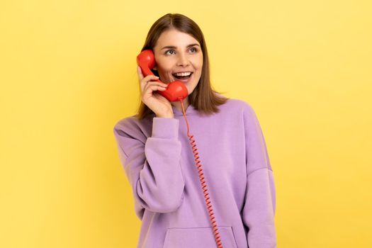 Portrait of beautiful young adult woman with excited expression talking on red retro telephone, holding handset, wearing purple hoodie. Indoor studio shot isolated on yellow background.