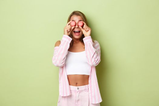 Portrait of silly blond girl playing with candies, holding macaroons over eyes and looking amazed, standing against green background.