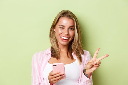 Close-up of attractive happy girl with blond hair, showing tongue and winking silly, make peace sign, using mobile phone, standing over green background.