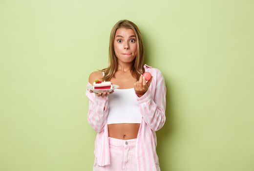 Image of attractive blond woman looking tempted and licking lips, holding macaroons and tasty cake, standing over green background.