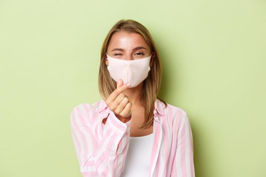 Concept of quarantine, coronavirus and lifestyle. Close-up of beautiful blond female model in face mask and pink shirt, showing heart sign, standing over green background.