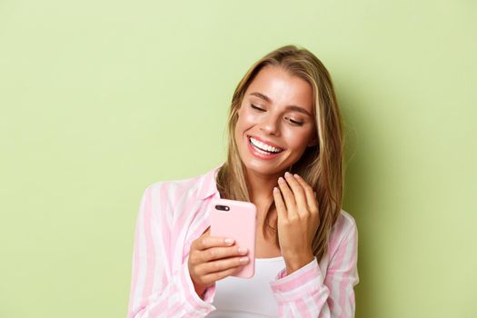 Close-up of attractive blond girl in pink shirt, messaging on phone, smiling and looking at smartphone, standing over green background.