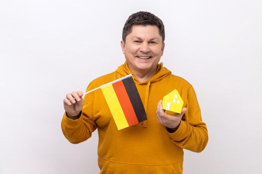 Optimistic man holding German flag and paper house, dreaming to buy his own apartrment in Germany, looking at camera, wearing urban style hoodie. Indoor studio shot isolated on white background.