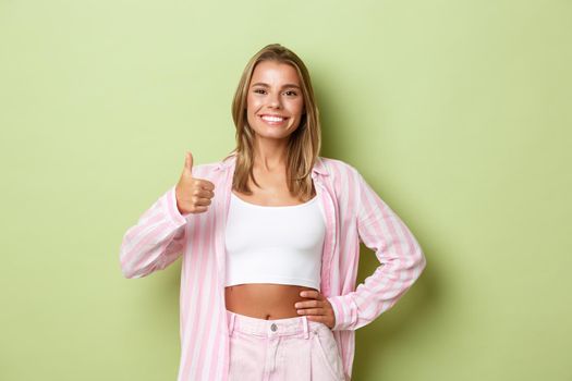 Image of confident modern woman with blond short hairstyle, wearing pink shirt and jeans, showing thumbs-up in approval, like and recommend something good, green background.