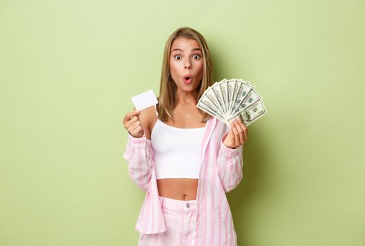 Image of impressed and excited blond woman, holding cash and credit card, standing over green background.