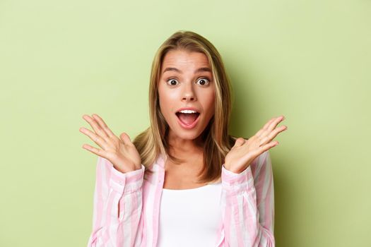 Close-up of attractive blond woman looking surprised, spread hands sideways and open mouth impressed, standing over green background.