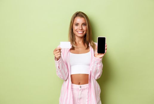 Image of stylish blond woman in pink shirt and jeans, showing smarpthone screen and credit card, recommending something online, standing over green background.
