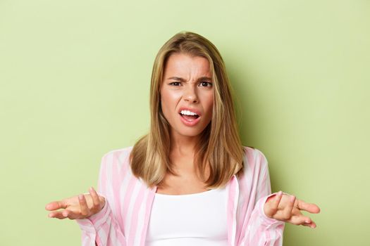 Close-up of pretty blond woman looking confused, raise hands sideways and arguing about something, standing over green background.