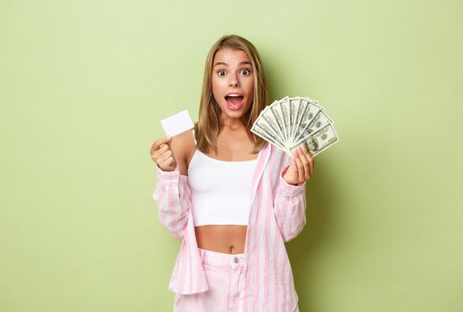 Portrait of excited blond woman showing credit card and cash, standing amazed over green background.