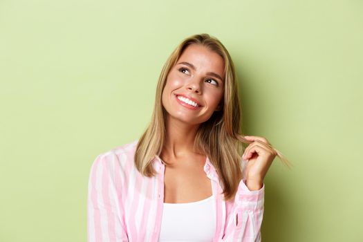 Close-up of glamour blond girl touching hair strand, smiling and looking dreamy at upper left corner, standing over green background.