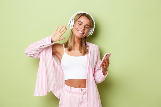 Image of pretty blond girl in pink shirt, listening music in headphones and smiling, holding mobile phone, standing over green background.