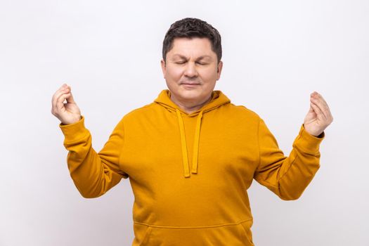 Portrait of handsome middle aged man standing with raised arms and doing yoga meditating exercise, mudra gesture, wearing urban style hoodie. Indoor studio shot isolated on white background.