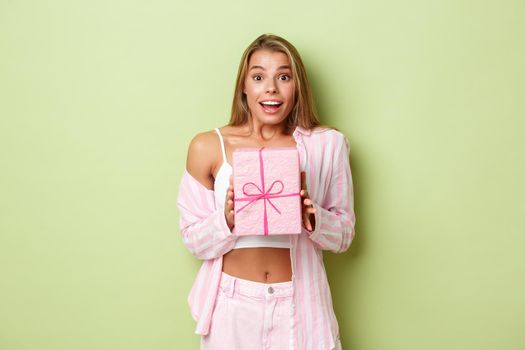 Portrait of attractive modern girl with blond short hair, looking excited, receiving a gift and smiling, standing over green background.
