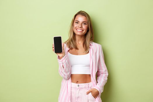 Image of smiling blond woman in pink striped shirt, showing mobile phone screen, standing over green background.