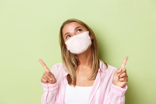 Concept of covid-19, social-distancing and lifestyle. Close-up of beautiful dreamy girl in face mask, looking and pointing up, standing over green background.