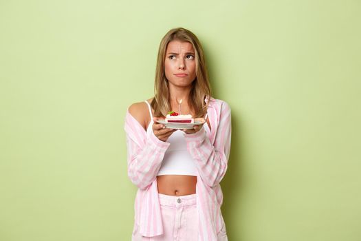 Image of gloomy blond girl with birthday cake, looking at upper left corner, puzzled to make wish, standing over green background.