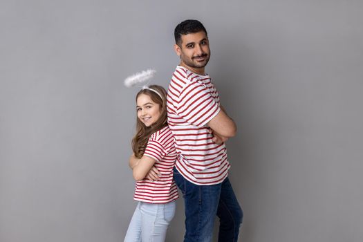 Portrait of lovely father and daughter in striped T-shirts, man and child with halo above head standing back to back with folded hands. Indoor studio shot isolated on gray background.