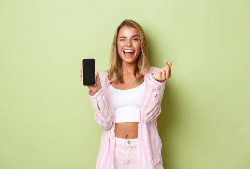 Image of stylish blond girl in pink shirt, showing heart sign and mobile phone screen, like something online, standing over green background.