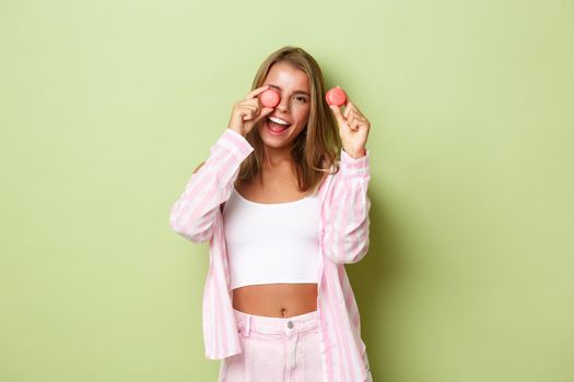 Portrait of beautiful modern woman with blond hair, showing pink macaroons and smiling, standing happy over green background.