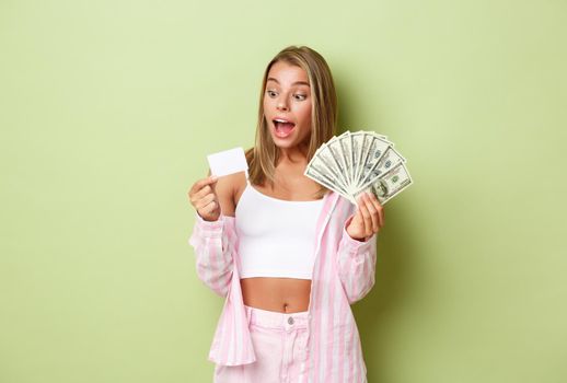 Portrait of attractive rich woman with blond hair, holding money and credit card and smiling amazed, standing over green background.