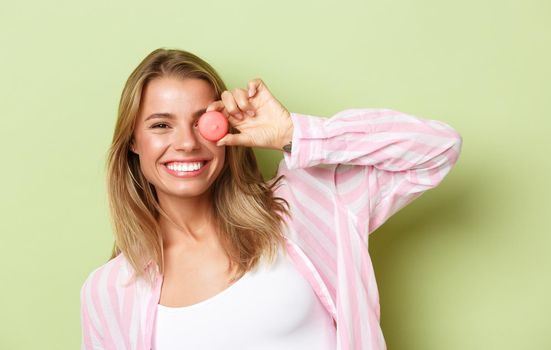 Close-up of playful smiling blond girl, holding macaroon near eye, standing happy over green background.