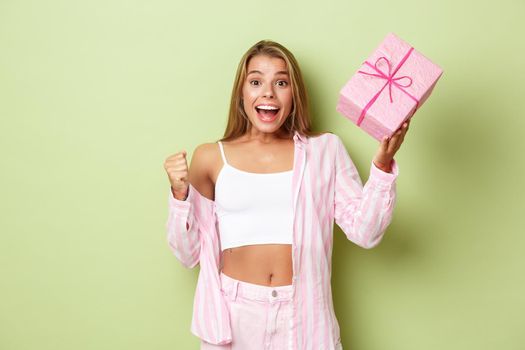Cheerful blond girl celebrating holiday, holding gift and looking excited, standing over green background.